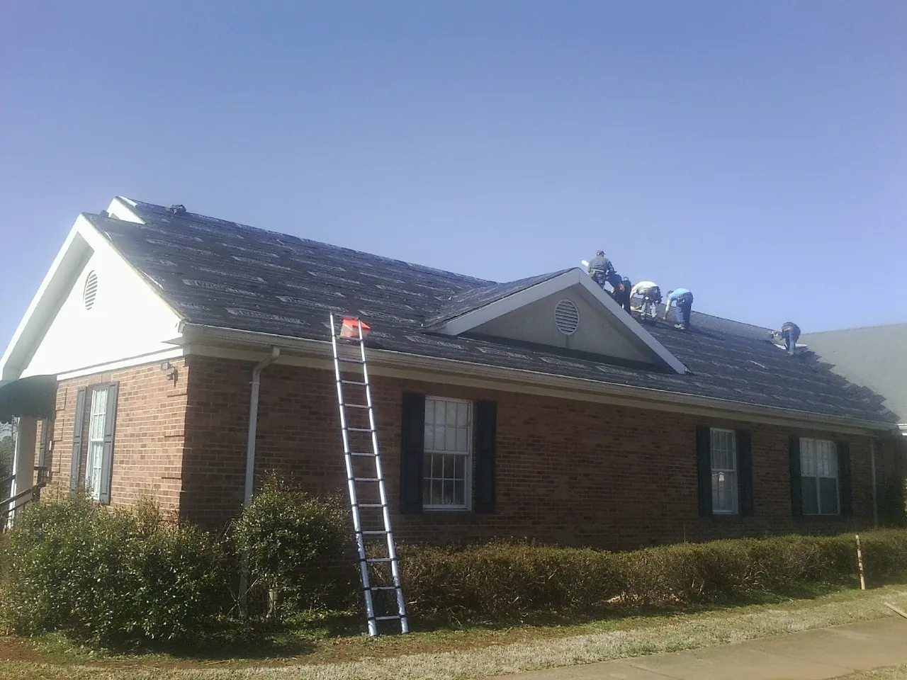 Crew tearing shingles off a brick ranch roof with a ladder set — Emergency Roof Repair work by our crew in Grimesland