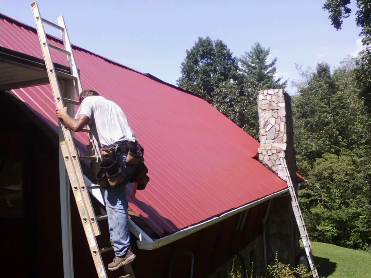 Roofer on a ladder beside a red metal roof and stone chimney — Soffit & Fascia Repair by Roofing Brothers in Grimesland